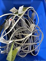 Blue storage bin filled with tangled white and black extension cords of varying lengths with attached tags and warning labels.