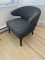 Side angled view of grey fabric arm chair showing curved backrest and tapered wooden legs on light wood floor.