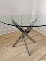 View of the round glass table showing the full glass top and criss-cross metal leg base on a light wooden floor