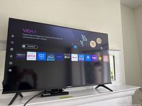 Front view of powered-on Hisense TV displaying VIDAA OS home screen with remote on stand
