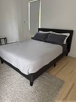 Full view of king size bed with tufted fabric headboard and matching frame in dark gray fabric, made up with white and dark gray bedding. The bed frame sits on wooden floor next to a window with a blind.
