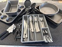 Photo showing various kitchen utensils, cutlery in tray, can opener, corkscrew, and baking pans including loaf pan, muffin tray, circular cake pans, and rectangular baking tray on countertop.