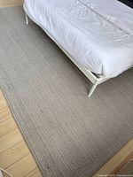 Photo showing the large area rug placed under a white bed frame on a wooden floor, highlighting the rug's size and neutral color.
