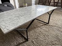 Side angled view of the marble coffee table on a textured rug showing the thin marble top and black metal legs with geometric design.