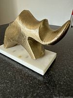 Angled view of the metal rhinoceros sculpture showing its textured golden surface and flowing horn shapes on a white marble base.