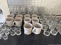 Full view of all items including 10 white ceramic mugs, 6 short clear glasses, 6 medium tall clear glasses, and 6 stemmed clear glasses arranged on a countertop.