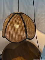 Pendant light with woven grid-patterned scalloped shade, lit and hanging over wooden surface, showing black cord and metal fitting
