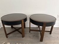 Pair of round side tables with wood legs, black leather apron, and glass tops placed on a light-colored wood floor.