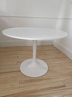 Full view of the white round pedestal dining table showing the table top and part of the base on a wood floor.