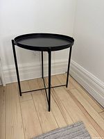 Full view of small black metal side table with removable tray top and tripod-style legs on wooden floor against wall