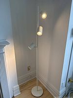 Full view of the white standing lamp placed in a corner near wall and floor molding, showing lamp base and three spotlight heads, all lit.