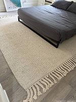 View of the woven cream area rug partially under a bed, showing chunky knit texture and fringed tassel edges.