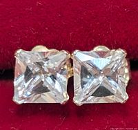 Front view of both square-cut cubic zirconia stud earrings showing clear sparkle and gold prong setting on red background.