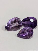 Three pear-shaped amethyst gemstones of varying purple shades, placed on a white background to show color, cut, and clarity.