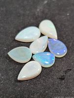 Seven pear-shaped white opal stones on dark background showing play-of-color in blue and green hues