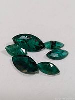 Photo of six marquise-cut genuine emerald gemstones on white background, showing color and size variation.