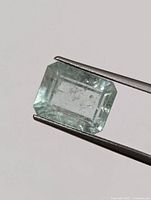 Close-up photo of a rectangular emerald cut green gemstone held by tweezers, showing internal inclusions and pale green color.