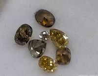 Close up of seven fancy color diamonds with various cuts, colors include yellow, brown, and grey tones on white background.