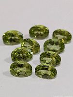 Nine oval-shaped genuine peridot gemstones arranged on a white background showing their facets and green coloration.