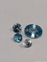 Four loose faceted gemstones showing three blue zircons and one clear zircon on plain background.
