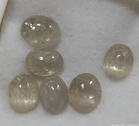 Six loose oval and round cabochon cut Zultanite gemstones laid on a soft white cloth background, showing transparent to slightly translucent clarity and natural inclusions with a pale color tone.