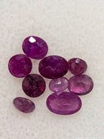 Nine faceted natural rubies on a white soft textured background, showing various sizes and deep pink to purplish red hues.