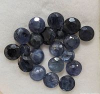Close-up photo of multiple round cut dark blue to blackish-blue sapphire gemstones laid on a white background showcasing their color and facets.