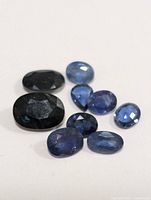 Clear image of nine loose genuine sapphire gemstones in various cuts and blue hues on a white background.