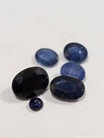 Six loose sapphire gemstones in assorted shapes and sizes on white background, showing color hues from dark to medium blue.