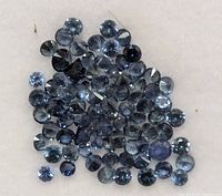 Close-up photo showing multiple small round blue sapphire stones on a white background, displaying facets and color.