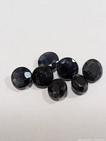 Photo showing a group of seven small, round faceted sapphires with dark blue-black color on a white background.