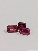 Three rectangular emerald-cut tourmaline gemstones, shades ranging from light to dark pink, placed on white surface.