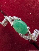 Close-up of the silver ring featuring an oval green emerald and side clear stones on a red velvet background.