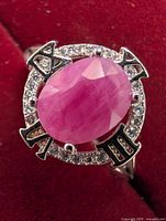 Close-up of silver ring featuring an oval faceted ruby surrounded by Roman numerals and small clear stones on a circular bezel.