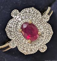 Close-up photo of a silver ring featuring an oval red ruby center gemstone surrounded by multiple small white stones arranged in floral design.
