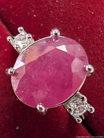 Close-up of ring top showing large oval ruby with two small round clear stones on each side in silver prong setting. Details of faceting and silver prongs visible.