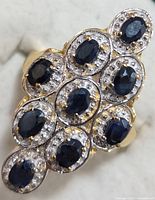 Top view of silver ring featuring nine oval blue sapphires with diamond halos in a cluster arrangement on a soft white background.