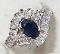 Close-up view of the sterling silver ring showing the oval blue sapphire center stone surrounded by baguette and round cubic zirconia stones in a swirl design.