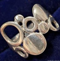 Close-up photo of silver ring showing the front and top side with circular and organic openwork shapes against a dark blue background.