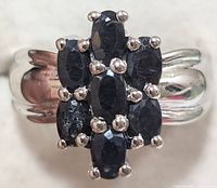 Close-up front view of silver ring with seven oval faceted dark blue sapphires set in prong settings clustered vertically on the polished silver band.
