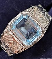 Close-up photo showing the rectangular emerald-cut blue topaz gemstone set in detailed silver with ornate textured patterns on the band.