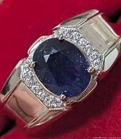 Top view of silver ring with large oval blue sapphire surrounded by small diamonds on a red fabric background.