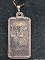 Front view of rectangular silver pendant showing blindfolded Lady Justice holding scales in raised relief