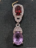 Front view of one silver earring with oval garnet top stone surrounded by cubic zirconia and dangling oval amethyst stone.