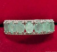 Close-up front view of silver ring with 9 oval emerald stones showing stone details and silver prong setting on red background.