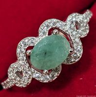 Close-up front view of silver ring with oval green emerald center stone surrounded by clear cubic zirconia stones on an ornate band.