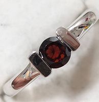 Close-up image of silver ring with a round garnet centerpiece and smooth polished band.
