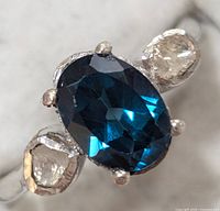 Close-up front view of silver ring with blue oval moissanite center and two clear diamond side stones.