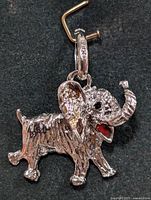 Close-up image of silver elephant pendant charm beside a brass hook, showing textured details and red enamel tongue.