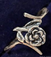 Close-up photo of silver ring with a rose floral design against a dark background showing details of petals and leaves.